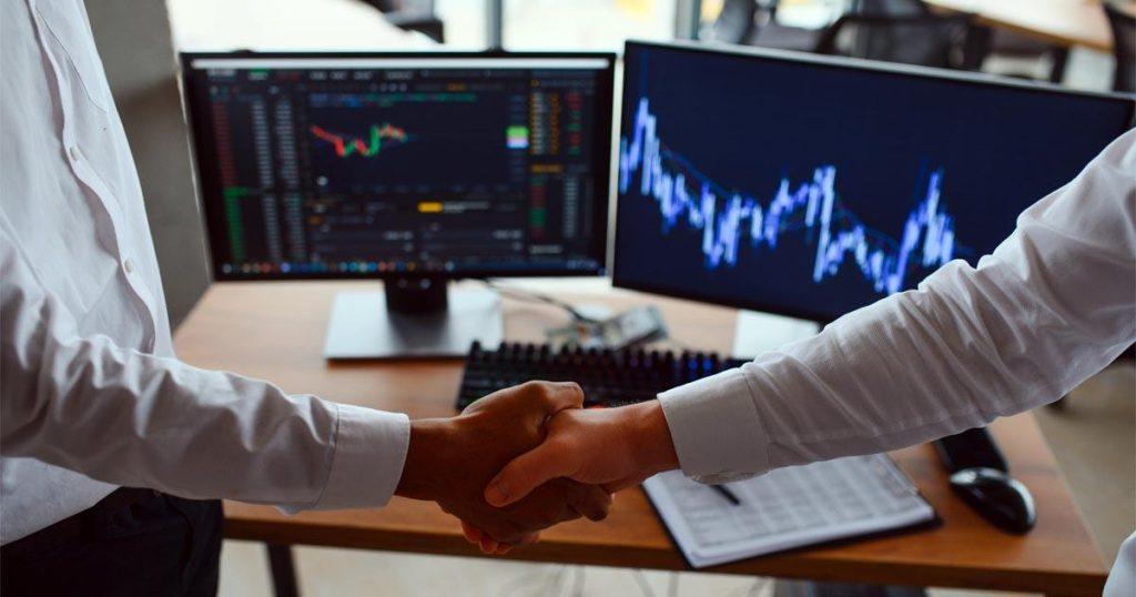 Duas telas de computador com gráficos de mercado financeiro e dois homens em frente apertando as mãos. Sobre o post "Curso Gratuito de Trading: vale a pena?".